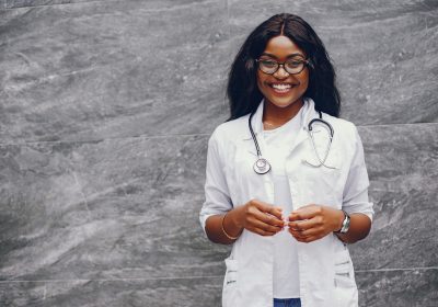 Stylish black doctor in a white uniform. Lady with stethoscope. Woman standing near gray wall